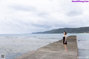 A woman in a black dress standing on a pier by the ocean.